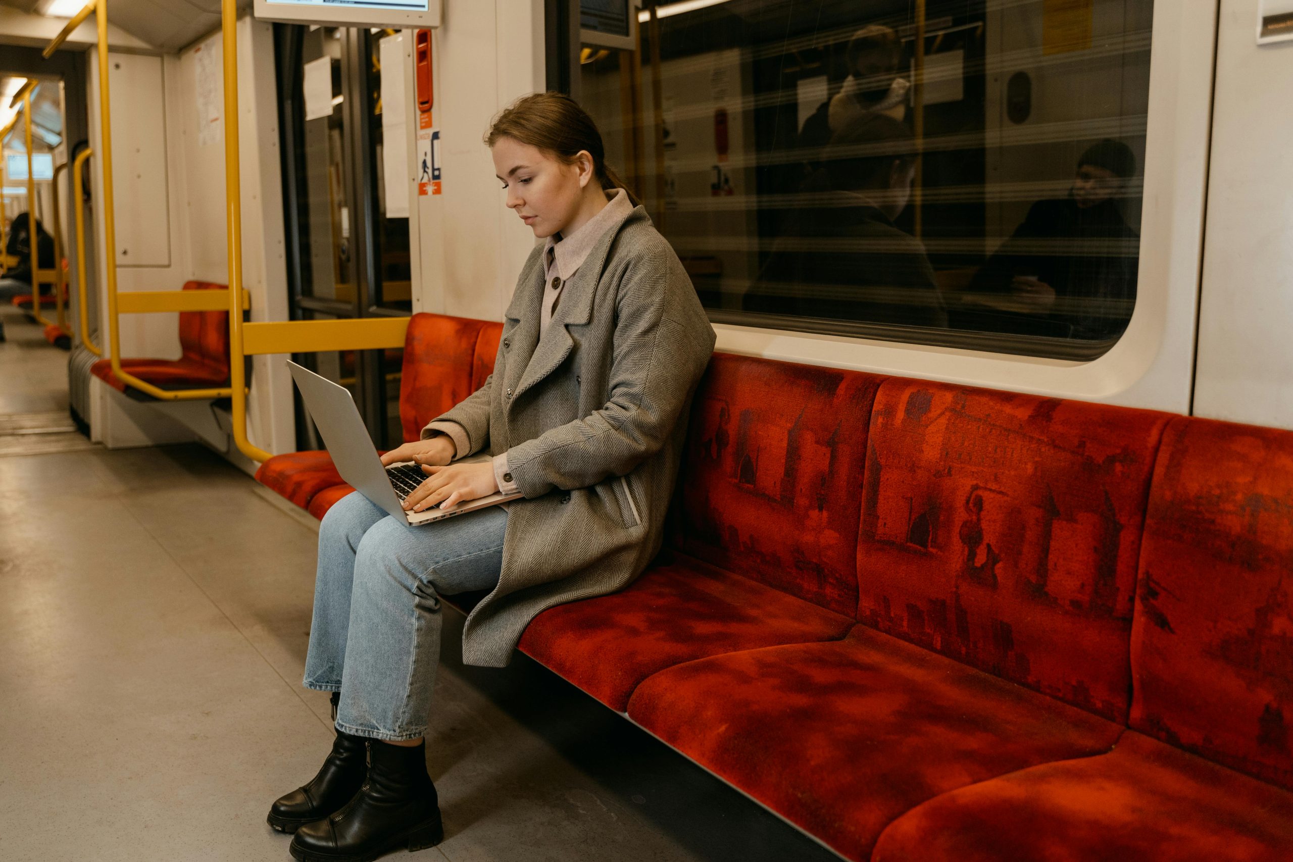 A woman in a gray coat works on a laptop while sitting in a subway car.